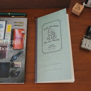 May include: A light green habit tracker notebook with the text "Trust The Seeds, You Are Planting". The notebook features a bear illustration and lines for daily and weekly tracking. A pen and other stationery items are visible.