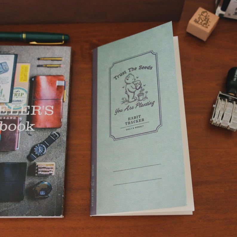 May include: A light green habit tracker notebook with the text "Trust The Seeds, You Are Planting". The notebook features a bear illustration and lines for daily and weekly tracking. A pen and other stationery items are visible.