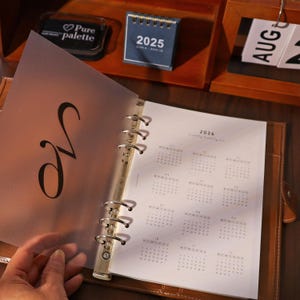 May include: A brown leather planner with a 2026 calendar page open. A clear overlay with a large, stylized letter "N" is in front. A small 2025 calendar and a "Pure palette" stamp are visible in the background.