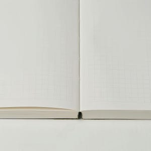 May include: Open notebook with grid-lined pages. The pages are white with a subtle grid pattern, perfect for writing or sketching. The notebook is open, revealing the inner pages and the binding.