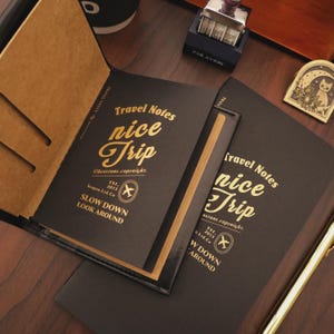 以下が含まれることがあります： 「Travel Notes nice Trip」と金色の文字が入った黒いトラベルジャーナル。ジャーナルは開いており、ベージュ色のページが見えます。「SLOW DOWN LOOK AROUND」というテキストも表示されています。スタンプと猫の形をした装飾が背景にあります。