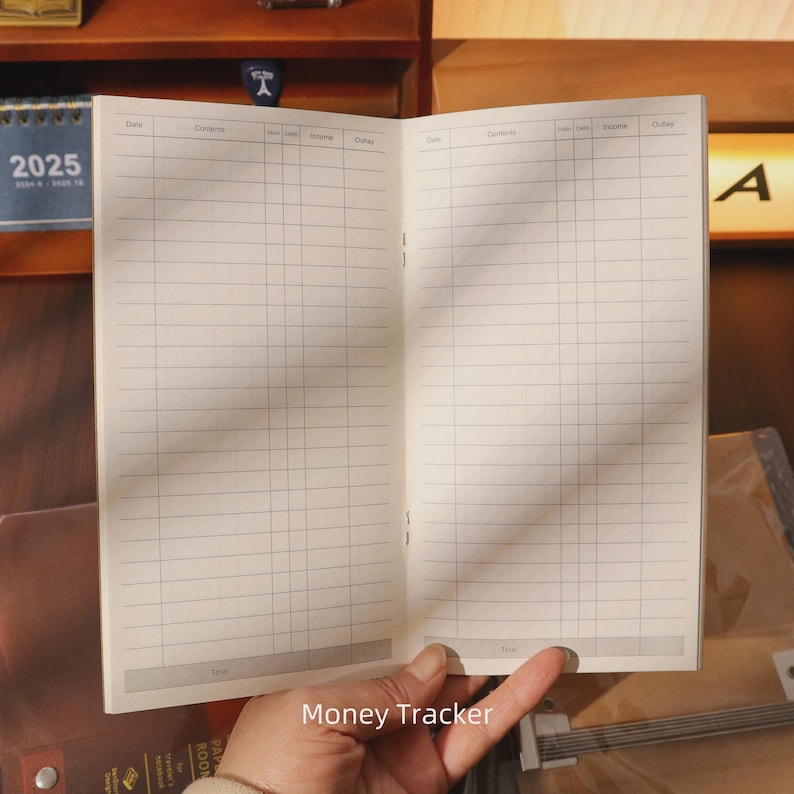 May include: Open notebook with lined pages, held open to display the interior. The notebook is labeled "Money Tracker" at the bottom. The pages are cream-colored, and the notebook is open to a double-page spread.