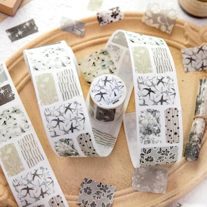 May include: Washi tape rolls and cut pieces with various patterns. The tape features designs of birds, flowers, and abstract shapes in shades of grey, white, and beige. The tape is displayed on a light brown tray.