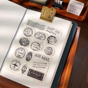 May include: A collection of clear stamps with black ink designs, including various city names, dates, and the words "AIR MAIL." A gold-colored book-shaped paper clip is also visible. The stamps are arranged on a white surface.