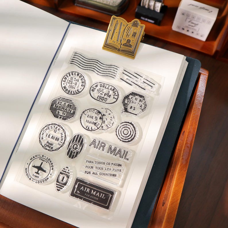 May include: A collection of clear stamps with black ink designs, including various city names, dates, and the words "AIR MAIL." A gold-colored book-shaped paper clip is also visible. The stamps are arranged on a white surface.