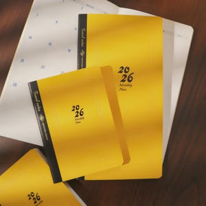 Puede incluir: Una colección de cuadernos amarillos de planificación mensual 2026 con lomos negros y el texto "Travel notes" y "BENITORE DESIGN". Los cuadernos están apilados sobre un planificador abierto blanco con un calendario.