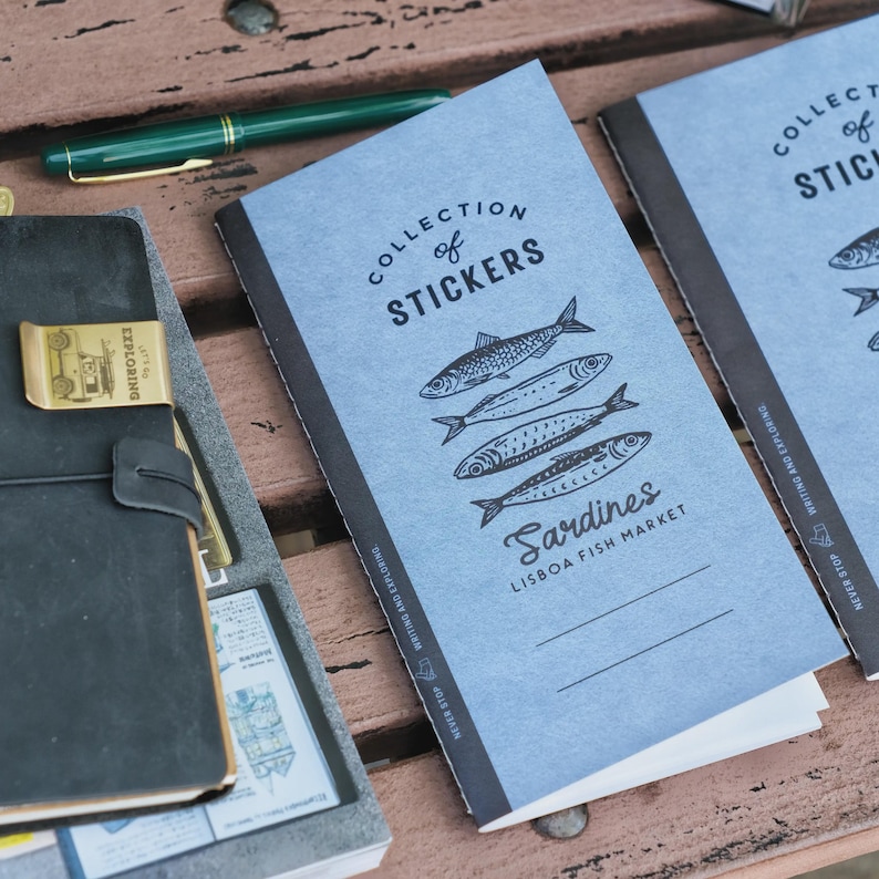 May include: A collection of stickers notebook with a light blue cover featuring illustrations of sardines. The notebook has the text "Collection of Stickers" and "Sardines Lisbon Fish Market". A green pen and a black leather journal are also visible.