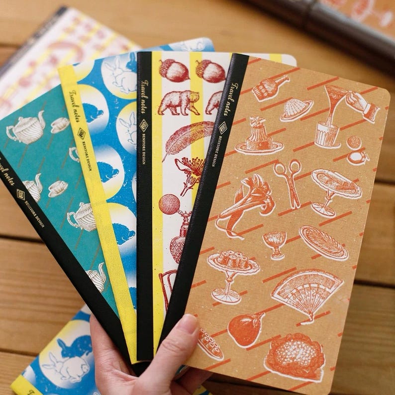 May include: A hand holds a stack of travel notebooks with various patterned covers. The notebooks feature illustrations of teapots, rabbits, acorns, bears, feathers, and other vintage-style designs. Each notebook has the text "Travel notes" and "BENTORE DESIGN" printed on the spine.