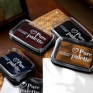 May include: A collection of rectangular ink stamp pads in various colors, including brown, black, and navy blue. Each pad is in a clear plastic case with the words "Pure palette" and "GLAY SHADES" printed on them.