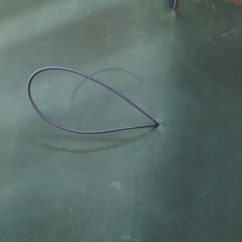 May include: A dark teal leather surface with a dark blue elastic loop. The loop is in the shape of a teardrop. The leather has a subtle texture and slight variations in color.