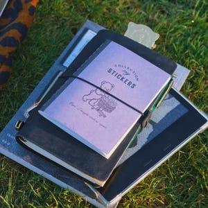 May include: A light purple notebook with a black elastic band featuring a line drawing of a bear pushing a shopping cart and the text "COLLECTION my STICKERS" and "Oops, I bought stickers again" resting on a black leather journal cover.