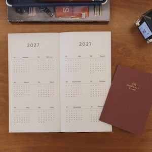 May include: An open 2027 calendar planner with cream-colored pages displaying twelve months, alongside a closed dark maroon notebook with the text The Monthly Plan embossed in gold on the cover, resting near a dark gray stationery box.
