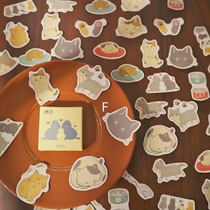 May include: A collection of cat-themed stickers in various poses and colors, including gray, yellow, and brown. Some stickers feature cats sleeping on plates. A small yellow box with cat illustrations is also present.