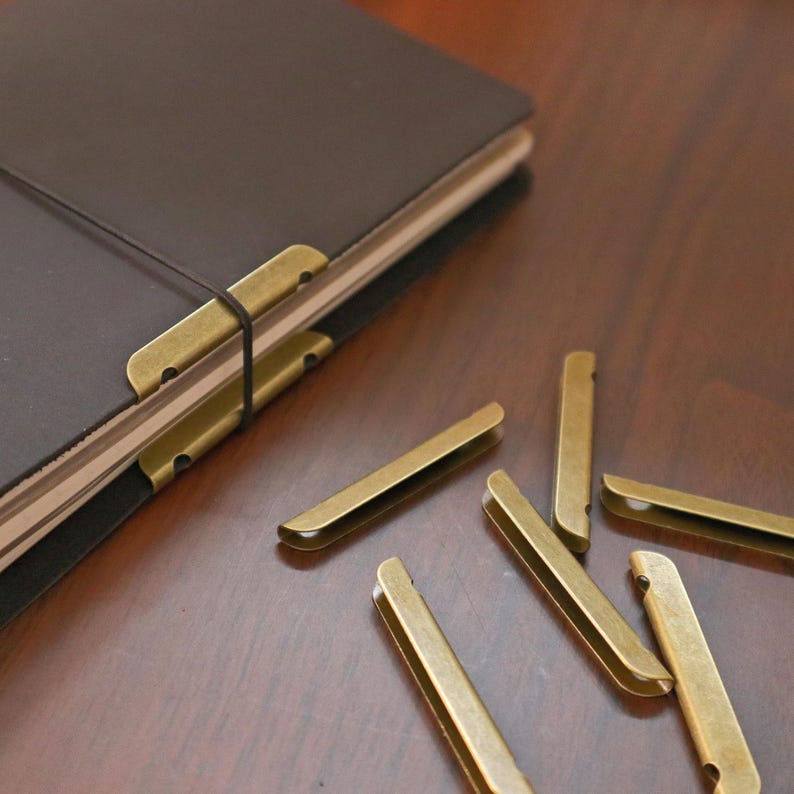 May include: A dark brown leather journal with brass-colored metal corner protectors and a matching elastic band closure. Several loose brass-colored metal corner protectors are scattered nearby.