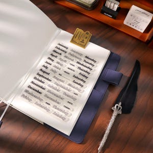 May include: An open notebook with a blue cover, featuring a clear sticker sheet with month and day labels. A gold-coloured book-shaped paper clip secures the sticker sheet. A decorative quill pen with a silver handle and black feather rests beside the notebook.