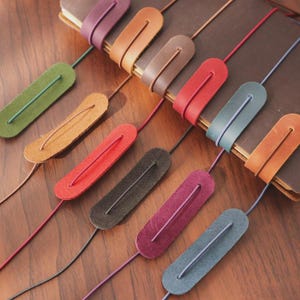 May include: A collection of leather bookmarks in various colors, including green, orange, red, and purple. Each bookmark features a leather loop and a thin cord, designed to be inserted into a book. The bookmarks are arranged on a wooden surface.