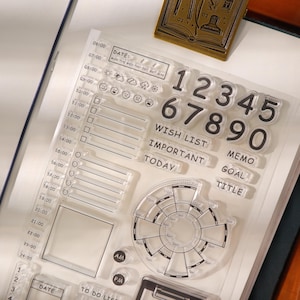 May include: Clear acrylic stamps for journaling and planning. Includes numbers, words like "Wish List" and "Today", and a circular time tracker. Also features date and to-do list stamps. Ideal for creating custom layouts.