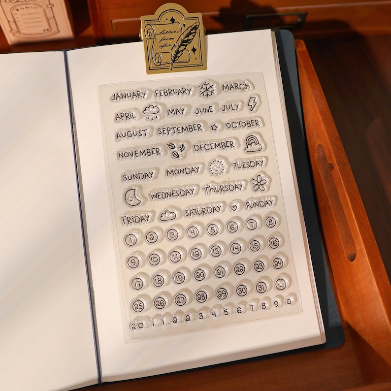 May include: Clear acrylic stamp set with months, days, and dates. Includes decorative icons and numbers. The stamp set is on a white background, held open by a gold-colored paper clip.