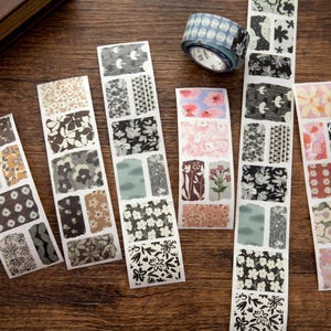 May include: A selection of decorative washi tape strips and a roll of tape featuring floral and geometric designs in brown, grey, pink, and blue. The strips are arranged on a wooden surface.