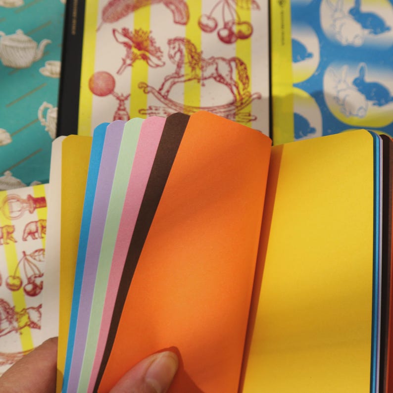May include: A collection of colorful notebooks with open pages. The pages are various colors, including orange, yellow, pink, and blue. The covers feature whimsical designs, such as a rocking horse, cherries, and rabbits.