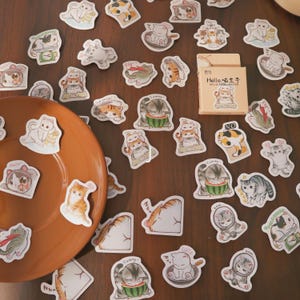 May include: A collection of cat-themed stickers scattered on a wooden surface, alongside a brown plate and a small box. The stickers feature various cat illustrations in different poses and colors, with some holding food or wearing accessories. The box has text on it.