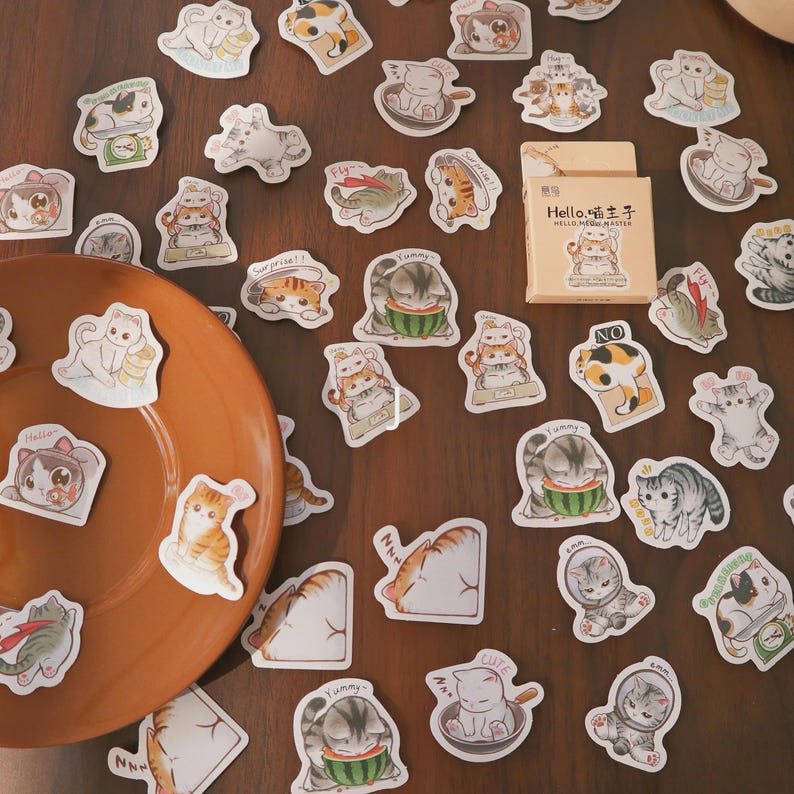 May include: A collection of cat-themed stickers scattered on a wooden surface, alongside a brown plate and a small box. The stickers feature various cat illustrations in different poses and colors, with some holding food or wearing accessories. The box has text on it.
