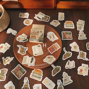 May include: A collection of vintage-style stickers scattered on a brown plate. The stickers feature illustrations of books, teacups, stationery, and a small box labeled "Cat Bookstore". The color palette includes warm tones of brown, beige, and cream.