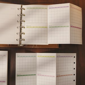 May include: Open planner with monthly calendar pages. The pages are white with a grid layout and colored headers for each month. The planner is bound with a metal ring binder and has a brown leather cover. The months are January, February, March, April, May, June, July, August, October, November, and December.