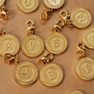 May include: Gold-colored initial charms with lobster clasps. Each charm features a raised letter within a circular, rope-edged frame. The letters visible are A, B, G, S, T, V, W, and Y. The charms are scattered on a brown surface.
