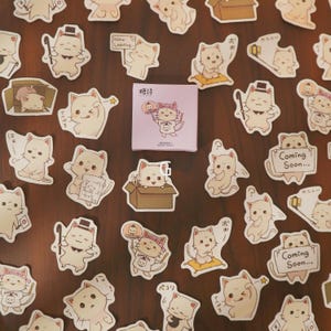 May include: A collection of cat-themed stickers and a small square box. The stickers feature various cartoon cats in different poses, some with accessories. The box has a cat illustration and Japanese text. The stickers are in shades of beige and white.