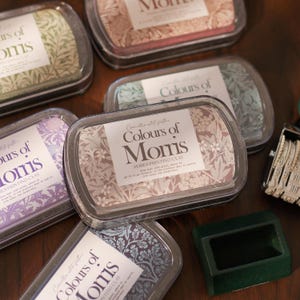 Könnte beinhalten: Mehrere rechteckige Behälter mit Drucktonerde mit floralen Designs und dem Text "Colours of Morris". Die Behälter sind transparent mit einem Kunststoffdeckel. Die Drucktonerde hat verschiedene Farben, darunter Rosa, Grün und Lila. Ein grünes Stempelkissen und ein Datumsstempel sind ebenfalls sichtbar.