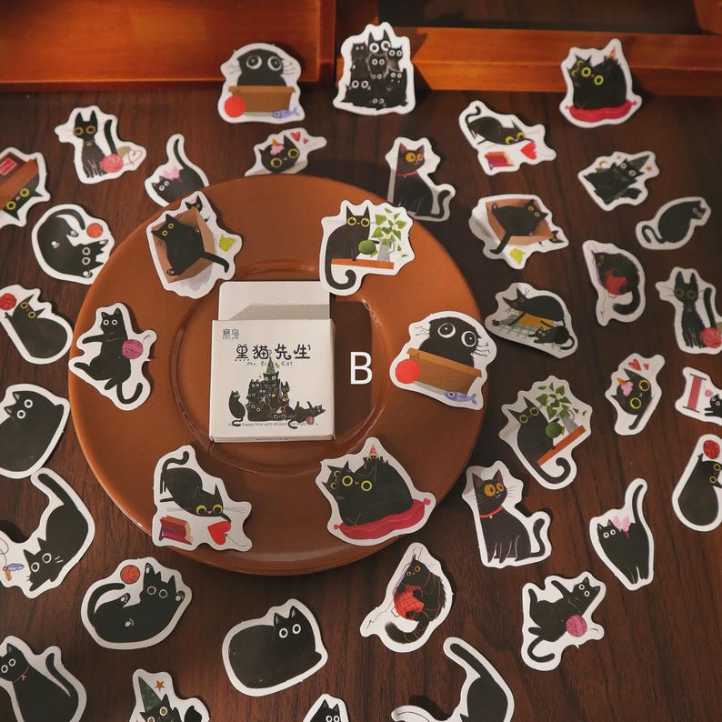 May include: A collection of black cat stickers in various poses, scattered on a brown plate and wooden surface. Some stickers feature cats with accessories. A small box with Chinese text and cat illustrations is also present.