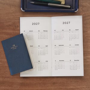 Könnte beinhalten: Ein dunkelblauer Planer mit dem Text The Monthly Plan in Goldfolie auf dem Einband, der auf einem offenen 2027-Kalender liegt. Der Kalender zeigt alle zwölf Monate des Jahres mit schwarzem Text auf weißen Seiten. Zwei Stifte liegen in einer blauen Ablage.
