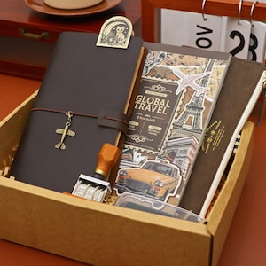 May include: A brown leather journal with a metal airplane charm and a decorative cat clip. The journal is in a cardboard box with a travel-themed sticker book, a brown zippered pouch, and a date stamp.