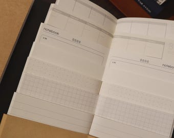 May include: An open notebook with white pages, featuring a weekly planner layout. The pages have a grid, dotted, and lined pattern. The notebook is partially inside a brown paper cover. The text "notebook" and "AM" are visible.