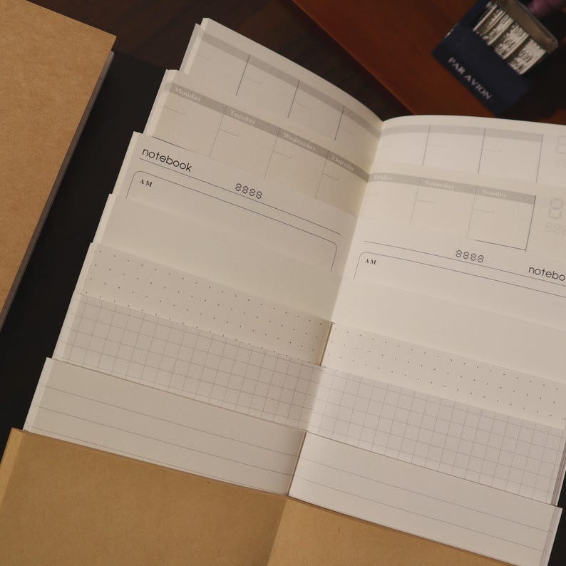 May include: An open notebook with white pages, featuring a weekly planner layout. The pages have a grid, dotted, and lined pattern. The notebook is partially inside a brown paper cover. The text "notebook" and "AM" are visible.