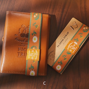 May include: A brown leather notebook with an elastic strap featuring a floral design and the text "NICI TRI". The elastic strap is orange with white daisies and green accents. A matching strap is on a cardboard sleeve.
