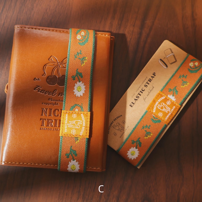 May include: A brown leather notebook with an elastic strap featuring a floral design and the text "NICI TRI". The elastic strap is orange with white daisies and green accents. A matching strap is on a cardboard sleeve.
