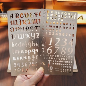 May include: Two metal stencils with cut-out letters and numbers. One stencil features uppercase and lowercase cursive letters and numbers. The other stencil has uppercase and lowercase block letters and numbers, with measurements of 6.35mm and 4.2mm.