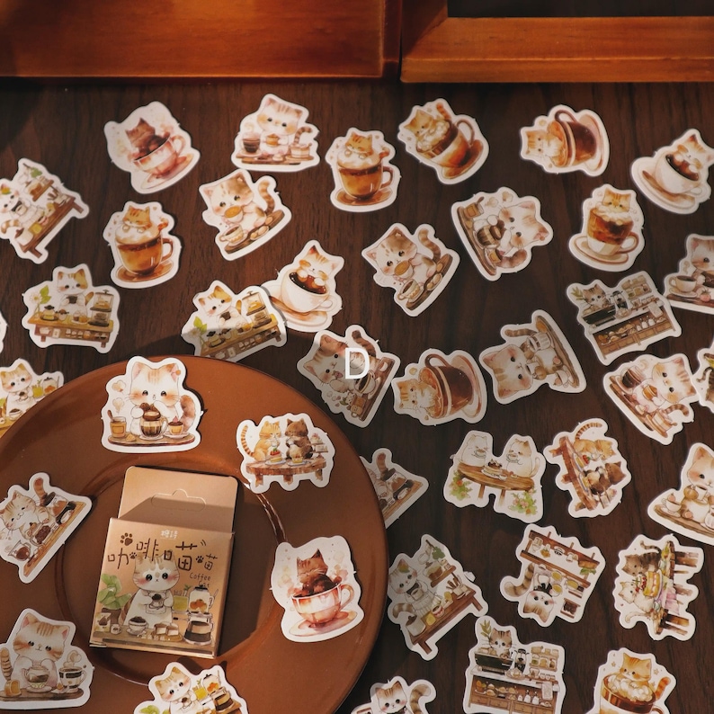 May include: A collection of cat-themed stickers featuring various designs, including cats with coffee cups, pastries, and cafe scenes. A small box of stickers is also visible, with Japanese text and a cat illustration. The stickers are in shades of brown, white, and beige.