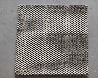 May include: Square black and white woven rug with a textured, dotted pattern. The rug is made of a durable material and is suitable for indoor use. The rug measures approximately 24 inches by 24 inches.