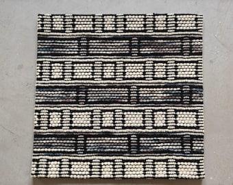 May include: Square, textured rug with a geometric pattern. The rug features rows of white, black, and gray beads, creating a striped design with rectangular accents. The rug is made of a thick, durable material.