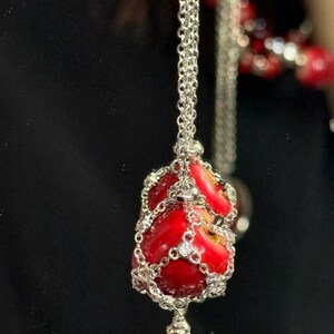May include: A silver necklace featuring a pendant shaped like a red apple. The apple pendant is encased in a silver filigree design and adorned with small, clear gemstones. The necklace hangs against a dark background.