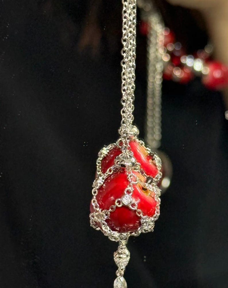May include: A silver necklace featuring a pendant shaped like a red apple. The apple pendant is encased in a silver filigree design and adorned with small, clear gemstones. The necklace hangs against a dark background.