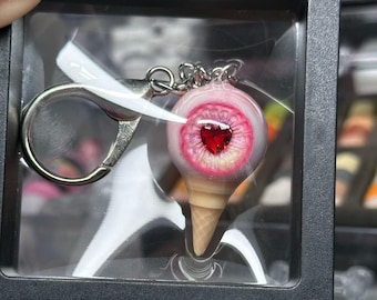 May include: A novelty keychain featuring a detailed eyeball design with a red heart-shaped pupil. The eye is set atop a miniature ice cream cone, with a silver-colored keyring attached. The keychain is displayed in a clear plastic case.
