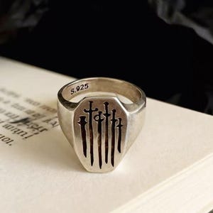 May include: A silver signet ring featuring an engraved design of five vertical swords on the face. The interior of the band is clearly stamped with the text S.925, indicating the metal composition of the piece. The ring has a smooth finish.