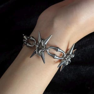 May include: A silver-colored bracelet featuring a series of pointed spikes and interconnected links. The bracelet has a gothic or punk aesthetic, with a bold and edgy design. The spikes are evenly spaced around the bracelet.