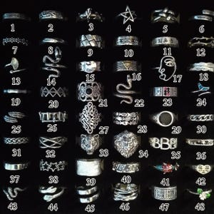 Pode incluir: Uma coleção de anéis prateados com vários designs, incluindo padrões geométricos, motivos de animais como cobras e leões, e símbolos como estrelas e rostos. Os anéis são exibidos em um fundo preto.