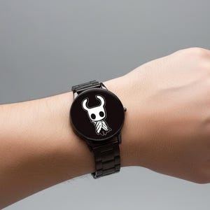 Puede incluir: Reloj de pulsera negro con un personaje de dibujos animados blanco en una esfera negra. El reloj tiene una correa de metal negro y una esfera redonda. El reloj se lleva en la mu&ntilde;eca sobre un fondo gris. Accesorio de moda.