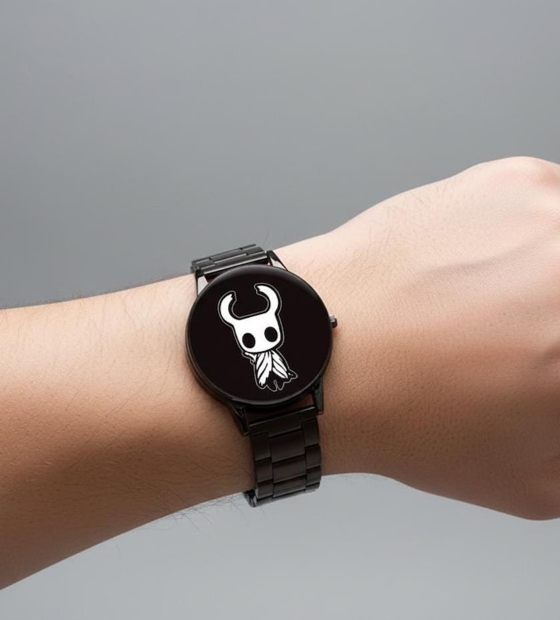 Puede incluir: Reloj de pulsera negro con un personaje de dibujos animados blanco en una esfera negra. El reloj tiene una correa de metal negro y una esfera redonda. El reloj se lleva en la mu&ntilde;eca sobre un fondo gris. Accesorio de moda.